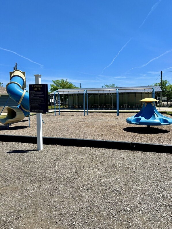 Ein Spielplatz mit Rutsche und Schaukeln, einem überdachten Sitzbereich und einem Schild mit Spielplatzregeln.