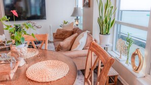 Ein gemütliches Wohnzimmer mit einem runden Holztisch, einer Couch und einem Fenster mit Pflanzen.