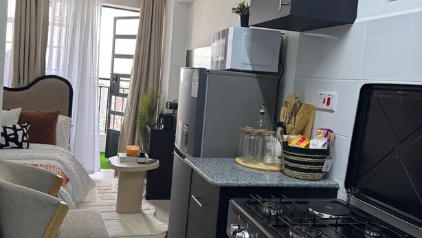 Et lille køkken med gasblus, køleskab og mikrobølgeovn. Der er et spisebord med stole.
