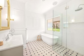 A bathroom with a white bathtub, a glass shower enclosure, a sink with a mirror, and a window with a view of greenery.