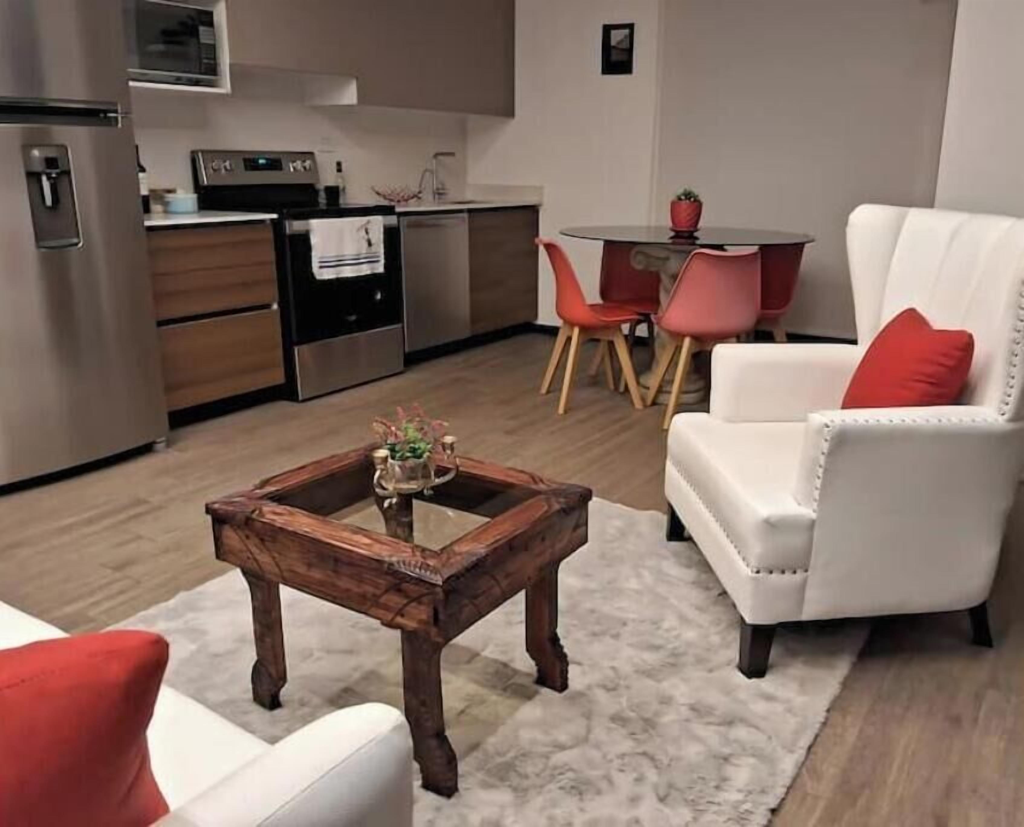 A modern kitchen with stainless steel appliances, a dining area with a round table and chairs, and a white armchair with a red cushion.