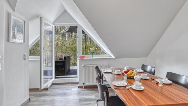 Een eetruimte met een houten tafel gedekt voor vier personen, een radiator en uitzicht op bomen door een raam.