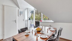 Een eetruimte met een houten tafel gedekt voor vier personen, een raam met uitzicht op bomen en een deur aan de linkerkant.