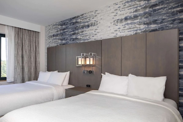 Habitación de hotel con dos camas, una pared de diseño de piedra, una mesita de noche con lámpara y una ventana con cortinas.
