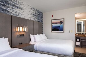 Habitación de hotel con dos camas, una mesita de noche con lámpara, una lámpara de pared, un cuadro enmarcado, un espejo y un baño pequeño.