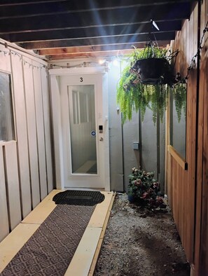 A narrow corridor with a door marked '3', a small step leading to a wooden deck, and a hanging planter with greenery. - Cozy 2 Bedrooms, Mountain View Private suite (Vernon)