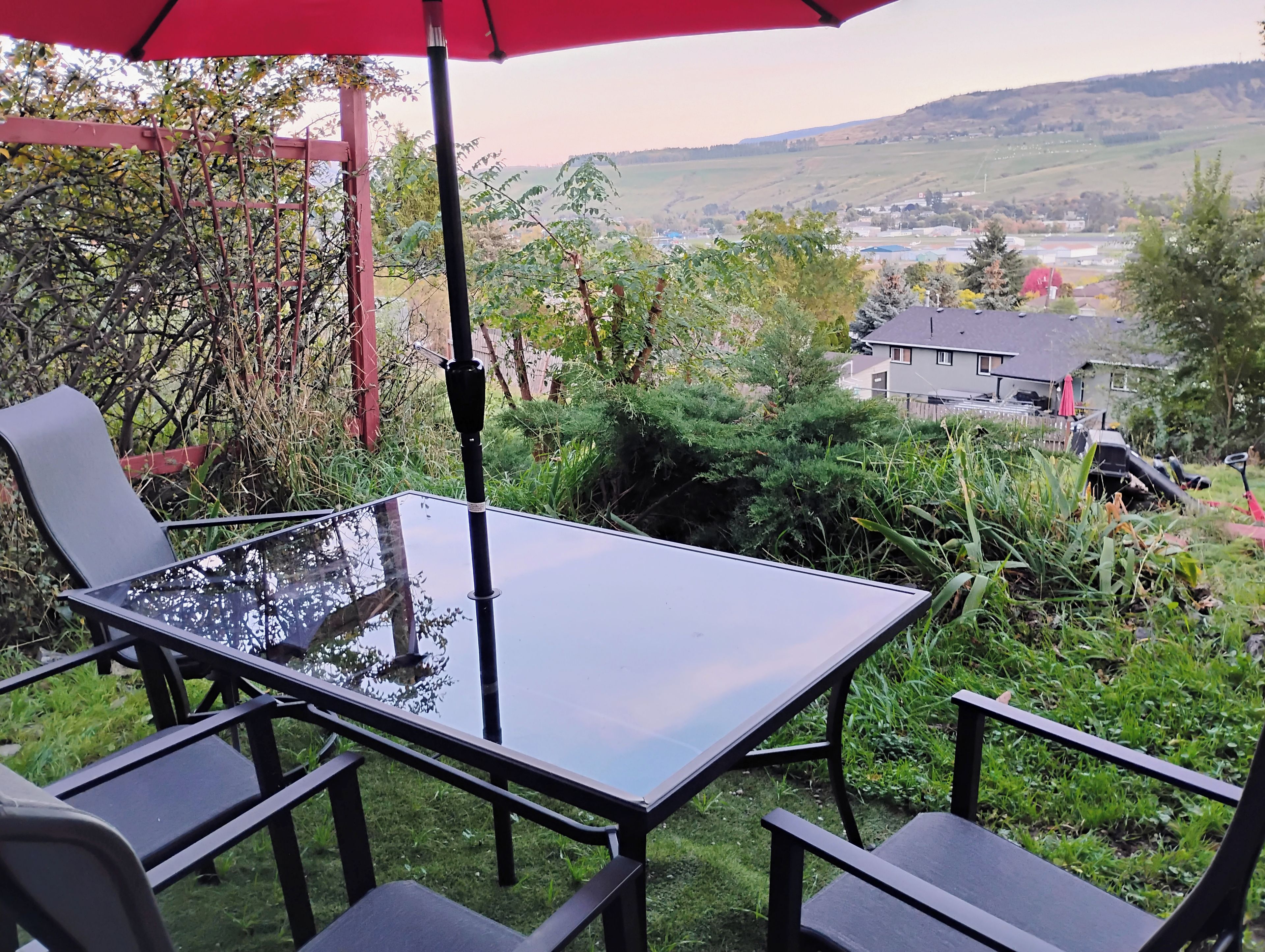 Un tavolo con piano in vetro, sedie, un ombrellone rosso e vista su un paesaggio rurale.