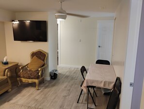 A living room with a television, a ceiling fan, a dining table with chairs, and a patterned armchair.