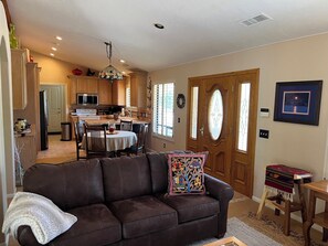 A living room with a brown leather sofa, a dining table with chairs, a kitchen in the background, and a wooden door with glass panels. - Peaceful 2-bedroom country house nestled on a 26 acre ranch in charming Ramona. (Ramona)