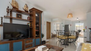 Een woonkamer met een televisie, houten planken, een eethoek met een tafel en stoelen, en een keuken op de achtergrond.
