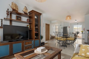 A living room with a TV, wooden shelves, a dining area with a table and chairs, and a kitchen in the background.