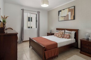 A bedroom with a bed, bedside tables, a nightstand, a lamp, a window with curtains, a plant, and a painting on the wall.