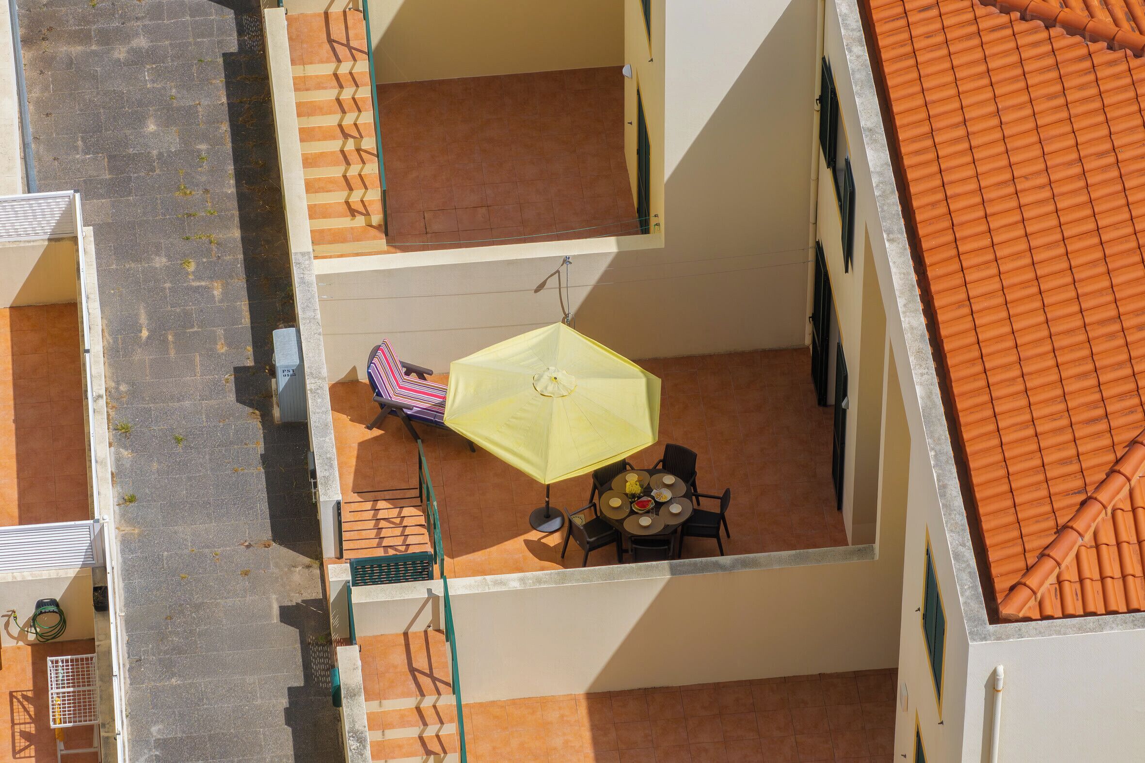 An outdoor balcony with a yellow umbrella, a round table with chairs, and a tiled roof.