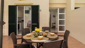Een terras met een tafel voor vier personen, omringd door rieten stoelen, onder een gele parasol.