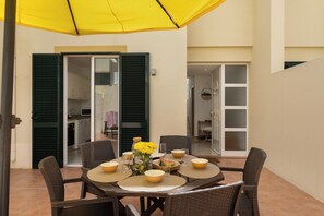 Un área de comedor al aire libre con una mesa preparada para cuatro personas, rodeada de sillas de mimbre, bajo un paraguas amarillo.