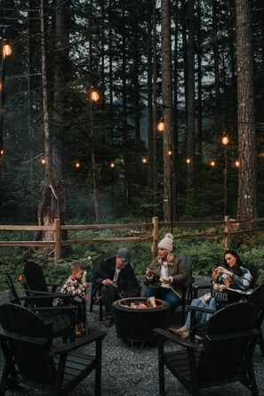 A group of people seated around a campfire in a forested area with string lights overhead. - Luxury Chalet • Infrared Sauna + Hot Tub • 10 min to Park (Ashford)