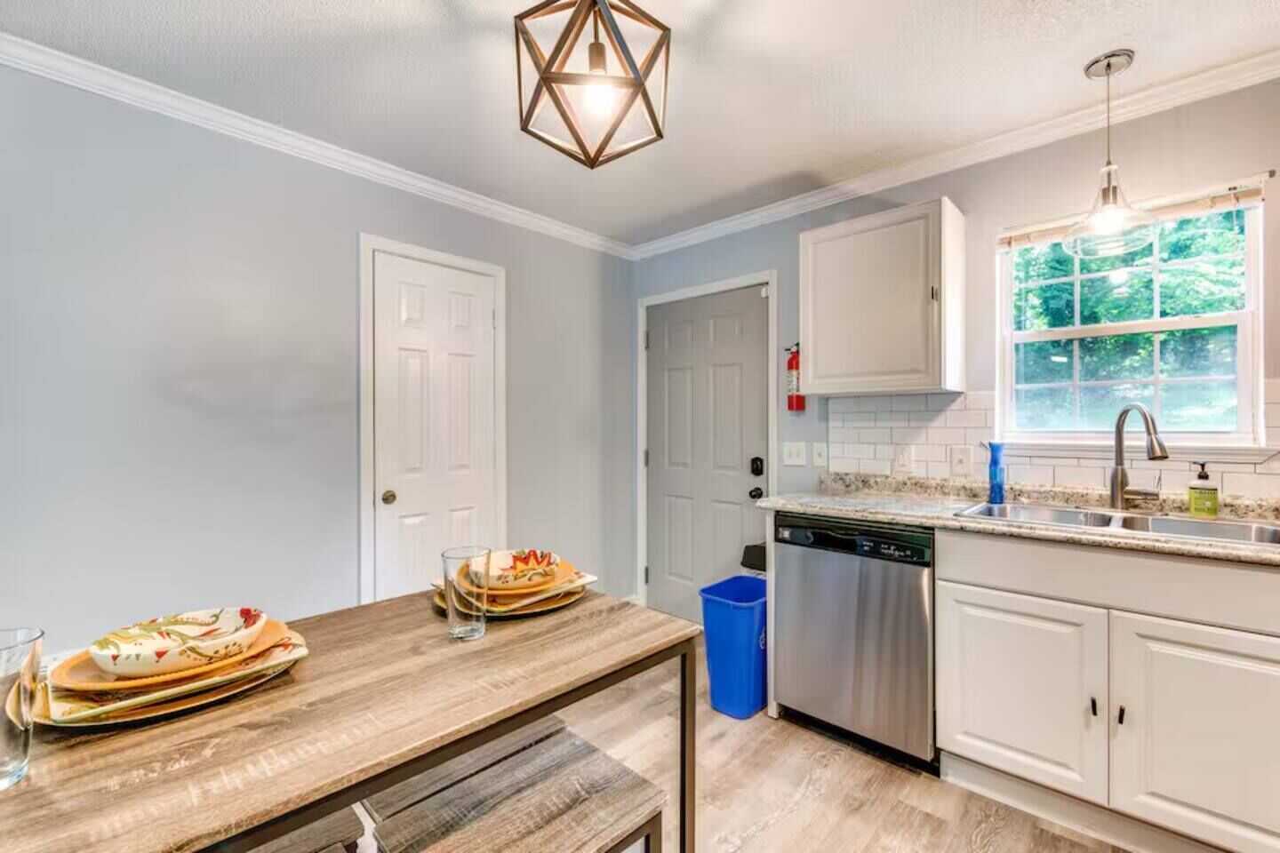 Una cocina con armarios blancos, lavavajillas, ventana con vista a follaje y mesa de madera con platos y un vaso.