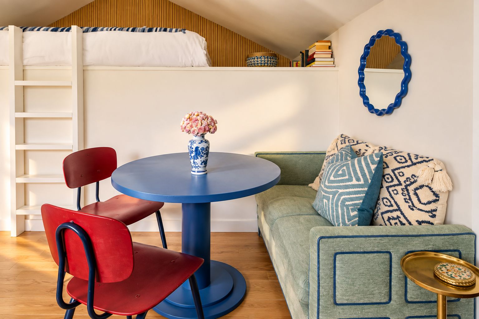 A room with a bunk bed, a round table with a vase, a red chair, a green sofa with patterned cushions, and a blue mirror on the wall.