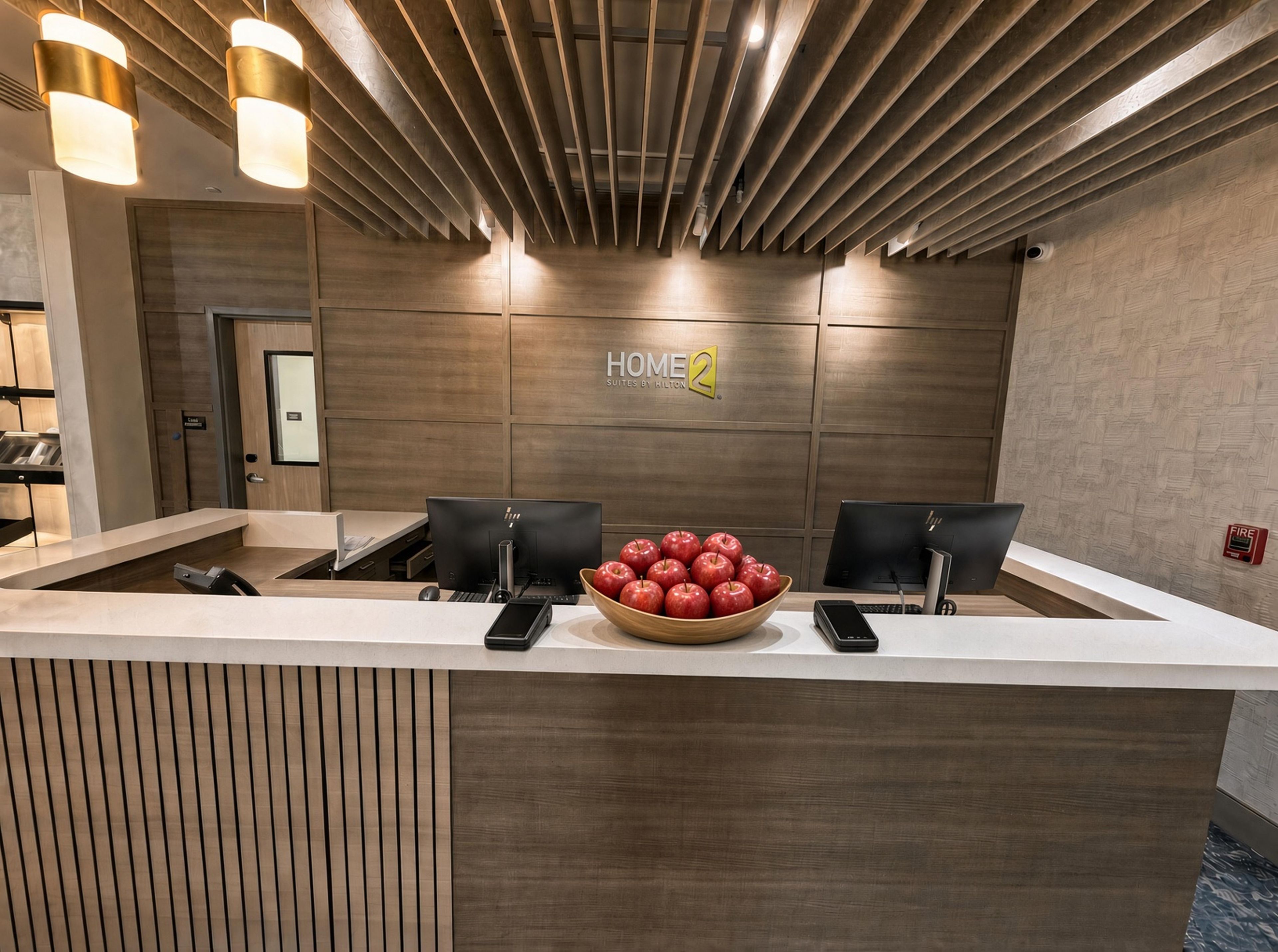 A reception desk with a wooden finish, two black chairs, a bowl of apples, and a 'HOME 2' sign on the wall.
