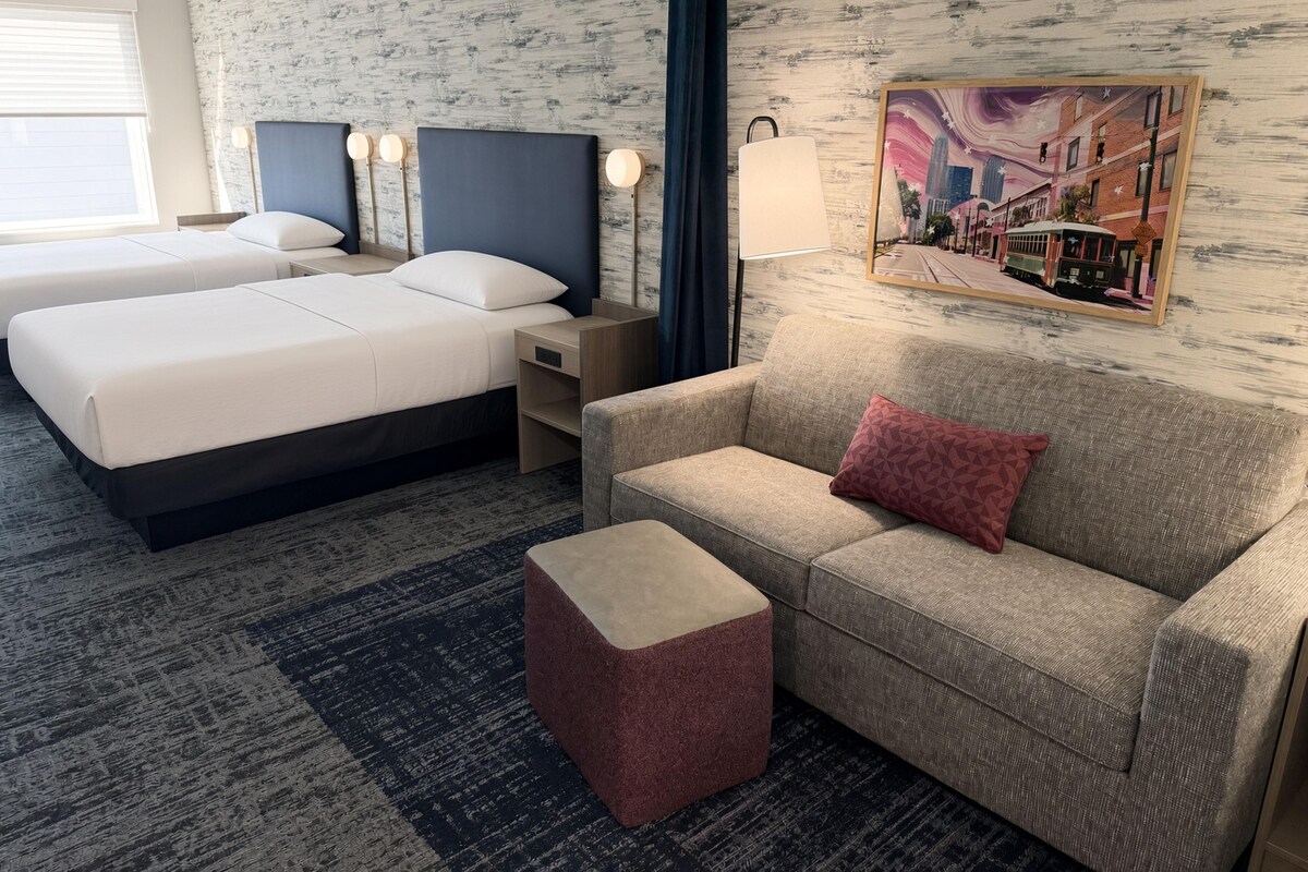 Quarto de hotel com duas camas, um sofá, um banquinho pequeno e uma luminária fixada na parede.