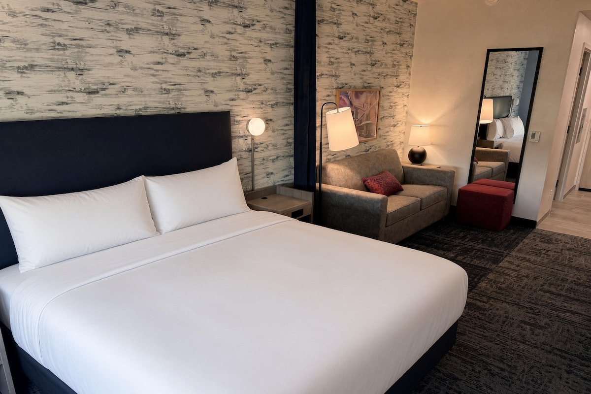 Quarto de hotel com uma cama grande, uma cadeira, um pequeno banquinho, uma mesa de cabeceira, um abajur e um espelho.
