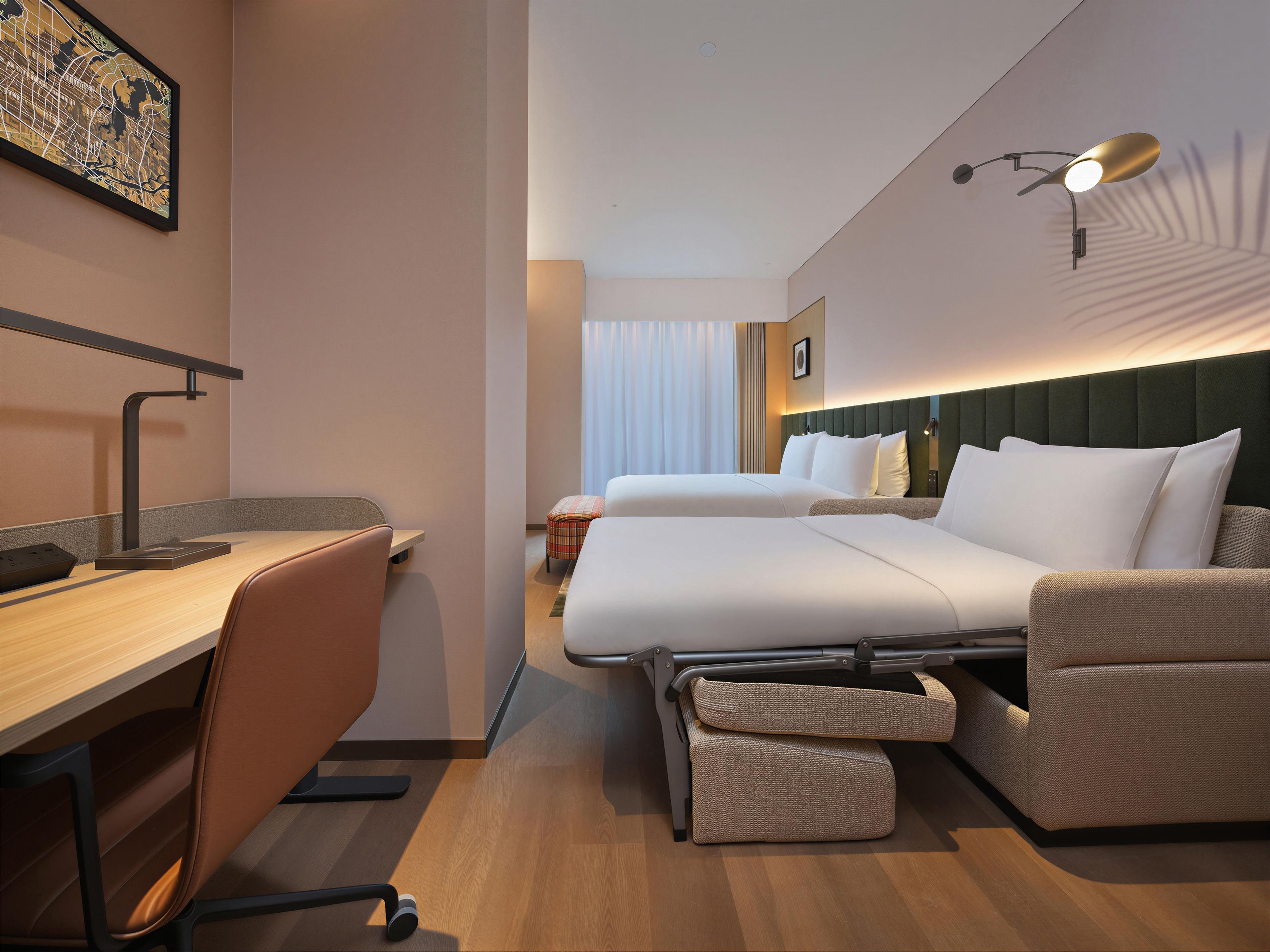 Una habitación de hotel con escritorio, dos camas, una silla y una lámpara fijada a la pared.