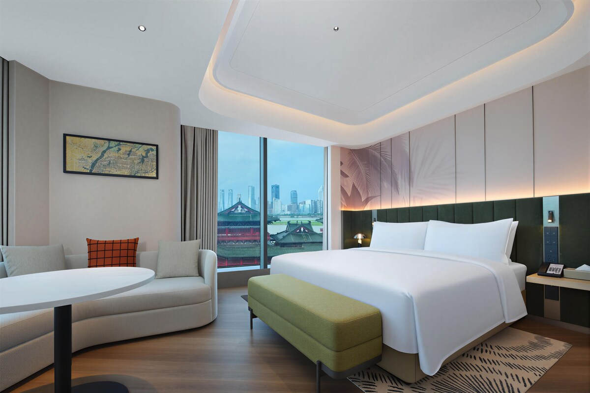 Une chambre d’hôtel moderne avec un grand lit, un canapé, une petite table et une vue sur le paysage urbain.