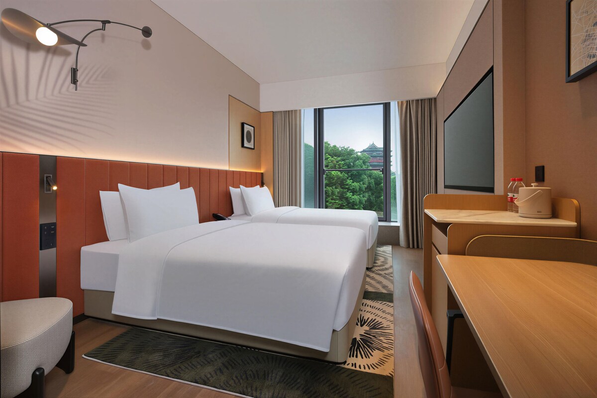 Une chambre d’hôtel avec un grand lit, un bureau, une chaise, une fenêtre donnant sur un espace verdoyant et un téléviseur fixé au mur.
