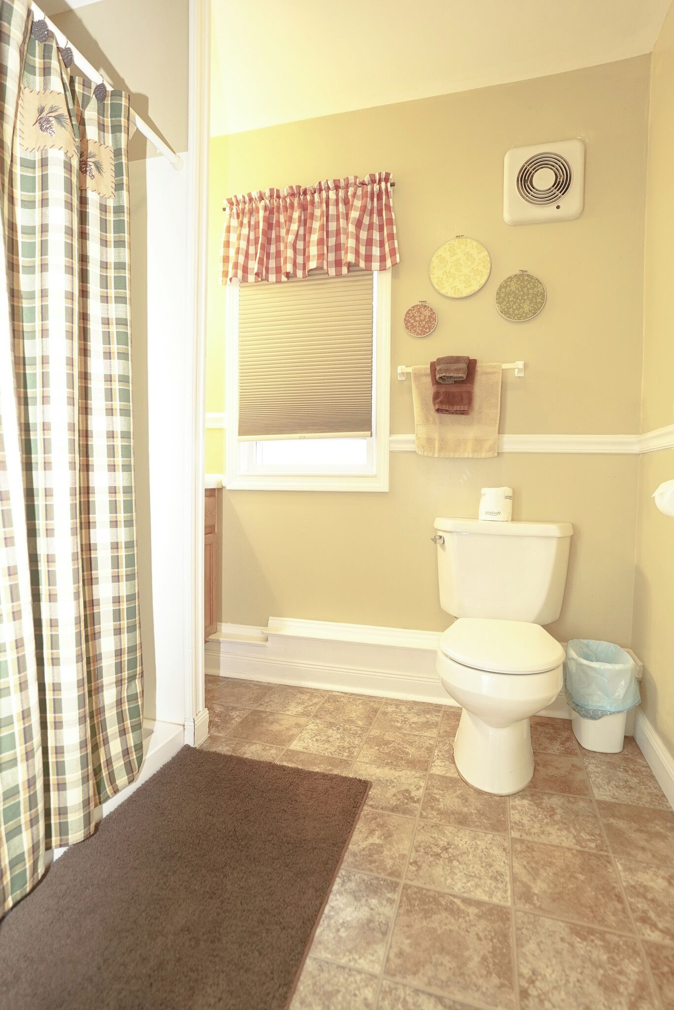 A bathroom with a toilet, a window with a checkered valance, a wall-mounted fan, and a towel rack.