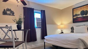 Un dormitorio con cama, una silla, una mesa, una planta en maceta, una ventana con cortinas, aire acondicionado, un cuadro y un ventilador de techo.