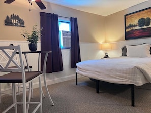 A bedroom with a bed, a chair, a table, a potted plant, a window with curtains, an air conditioner, a painting, and a ceiling fan. - Beartooth Suites (Joliet)