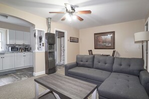 A living room with a gray sectional sofa, a wooden coffee table, a ceiling fan, and a kitchen in the background. - Beartooth Suites (Joliet)