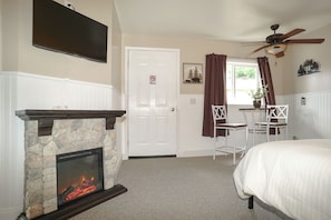 A bedroom with a stone fireplace, a bed, a desk with two chairs, a ceiling fan, and a window with curtains.