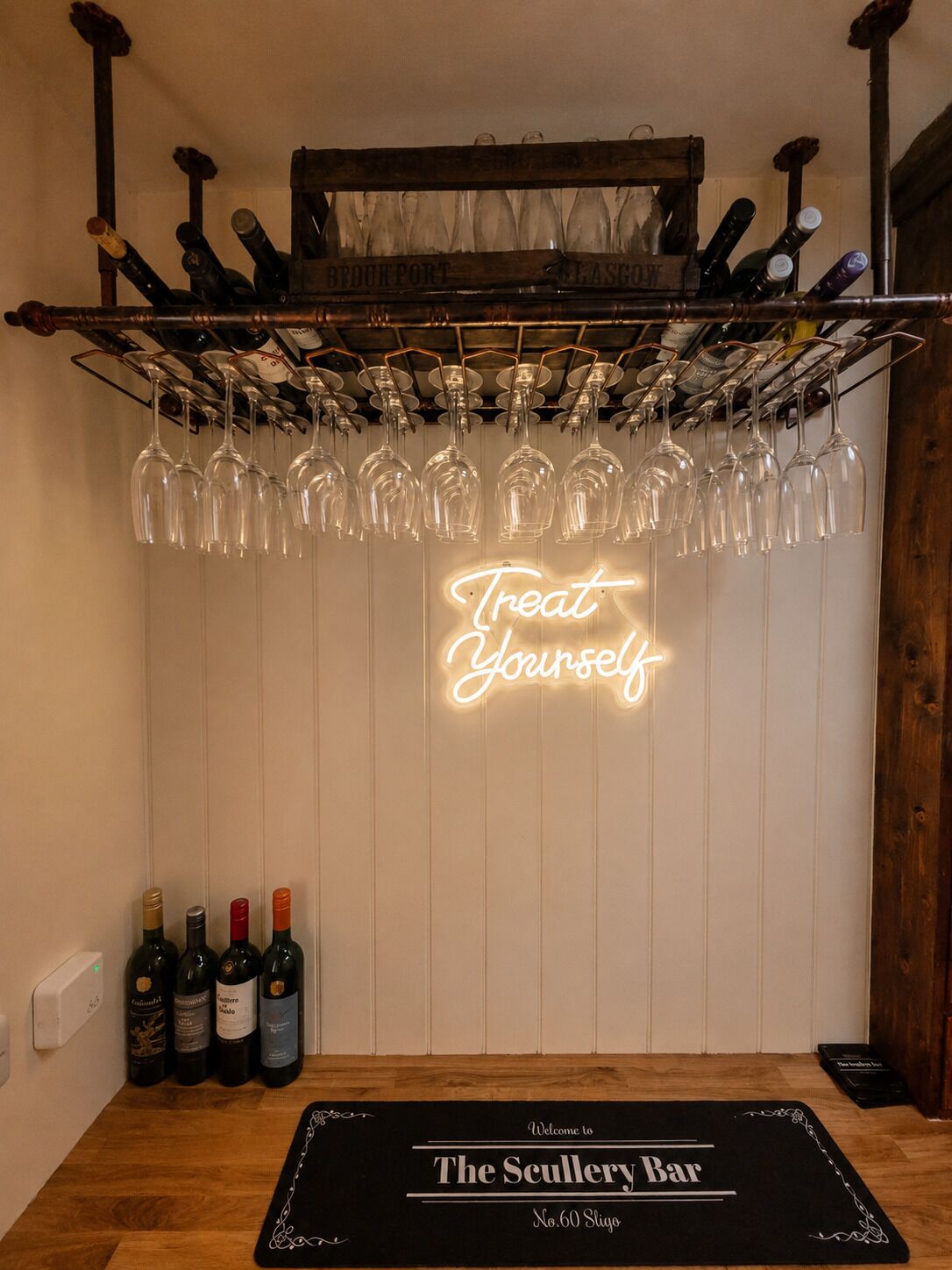 A bar area with a neon sign reading 'Treat Yourself,' wine bottles, and a welcome mat for The Scullery Bar.