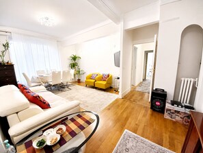 Ein modernes Wohnzimmer mit Sofa, Couchtisch und einem gelben Sessel. Es gibt einen Essbereich mit Tisch und Stühlen sowie eine Küchenzeile mit Geräten.