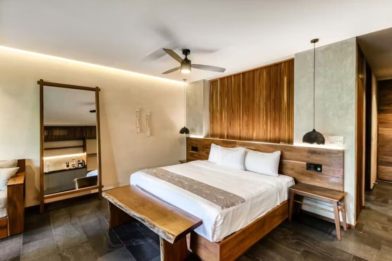 A modern bedroom with a bed, bedside tables, a bench, a mirror, a ceiling fan, and a wooden panel wall.