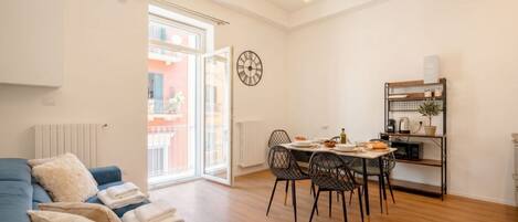 A modern living room with a sofa, dining table, chairs, a clock, and a kitchen area with a shelf.