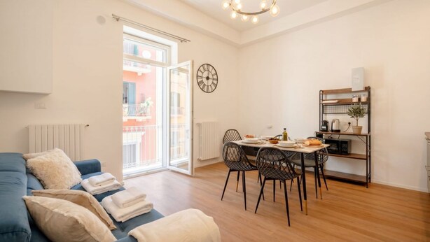 En moderne stue med sofa, spisebord, stole, et ur og et køkkenområde med en hylde.