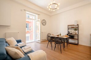 Ein modernes Wohnzimmer mit Sofa, Esstisch, Stühlen, einer Uhr und einer Küchenzeile mit Regalen.