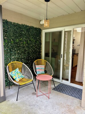 A balcony with two wicker chairs, one with a colorful cushion and the other with a striped cushion, a small pink table, and a green leafy wall.
