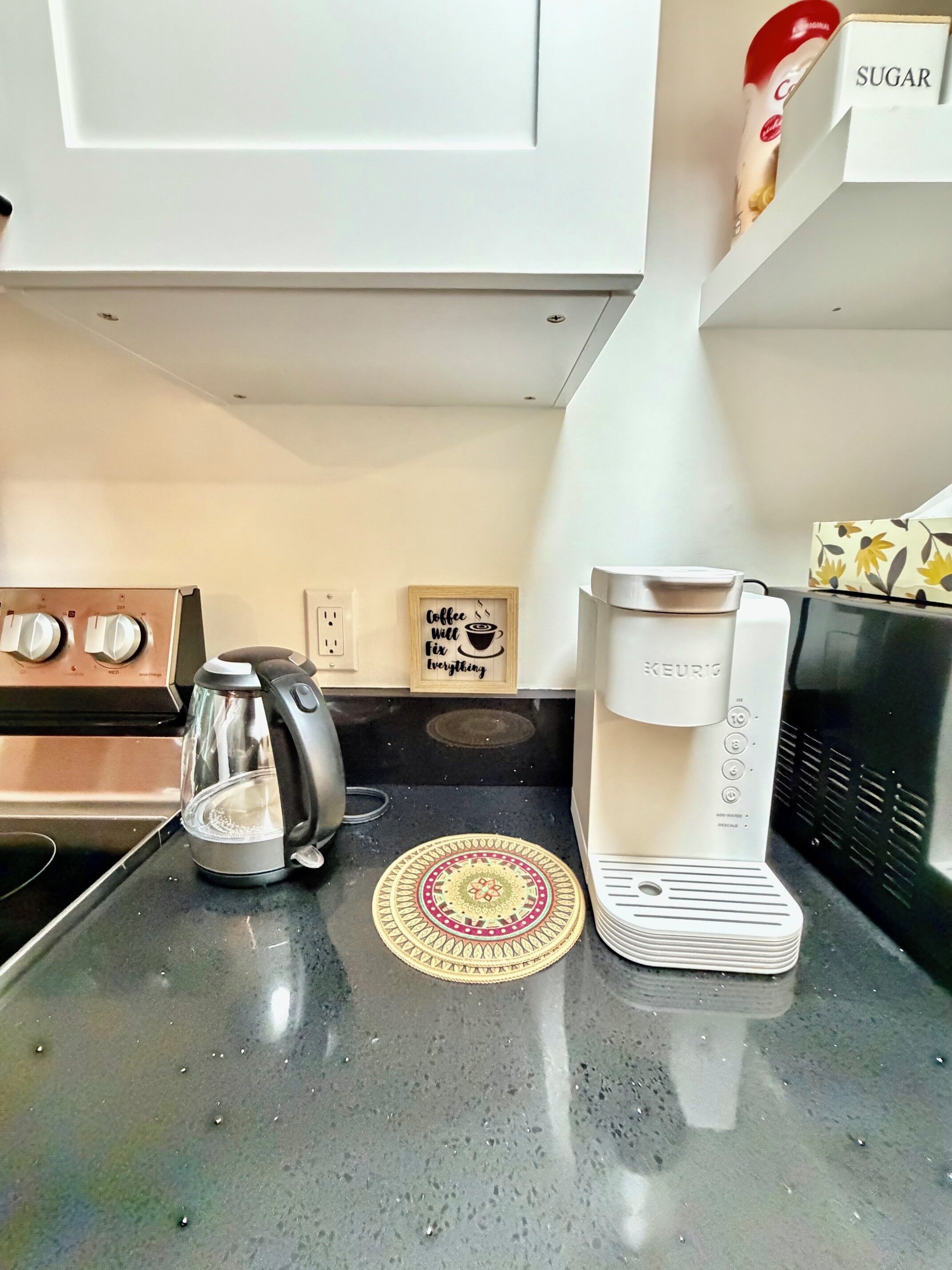 Keittiössä on Keurig-kahvinkeitin, lasinen kahvipannu ja sokeripurkki työtasolla.