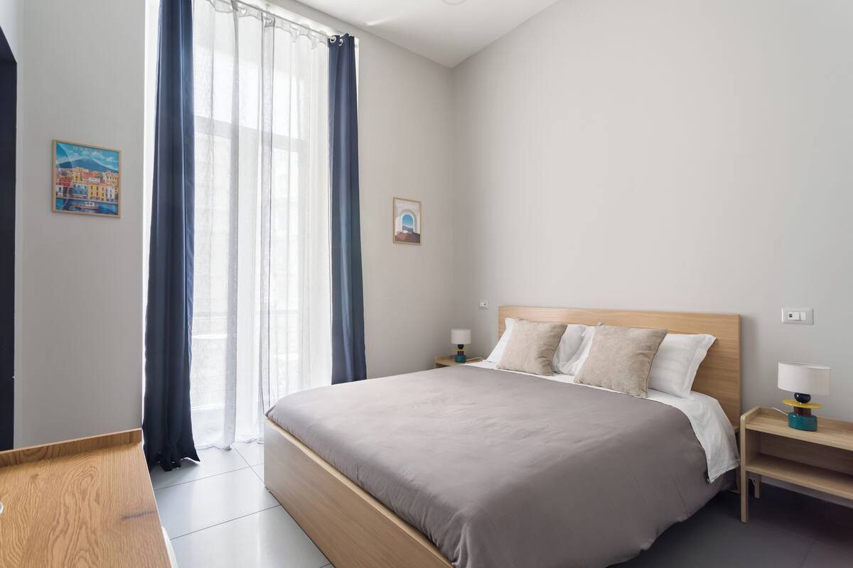 A modern hotel room with a large bed, bedside tables, and a window with curtains.