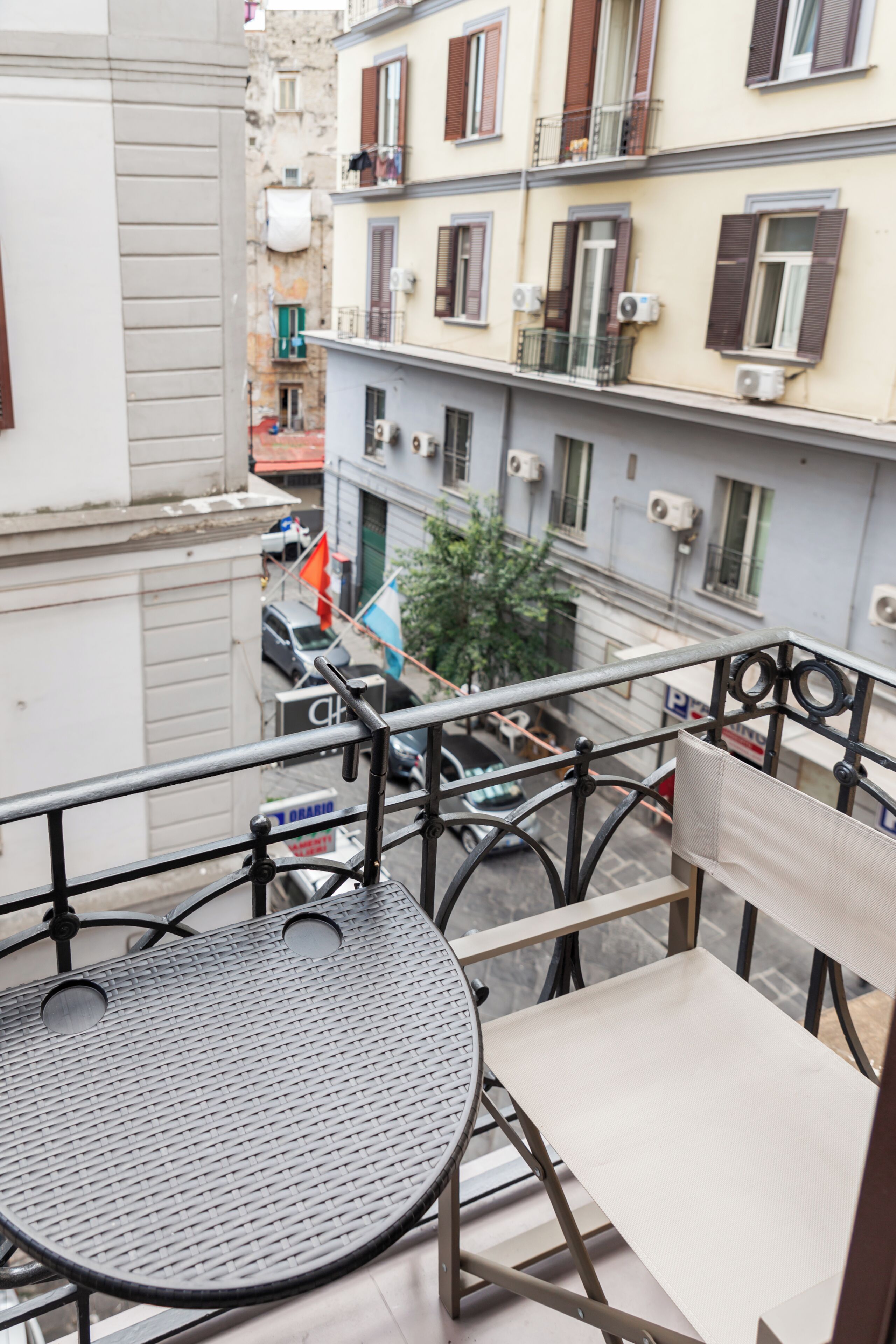 A balcony with a metal table and chair, overlooking a street with parked cars and buildings.