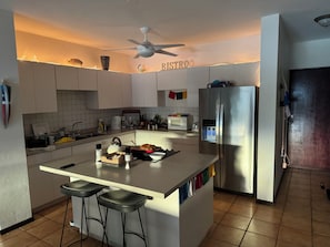 A modern kitchen with a central island, bar stools, a refrigerator, a microwave, and a ceiling fan. - Ocean Sunset Lover (Isabela)