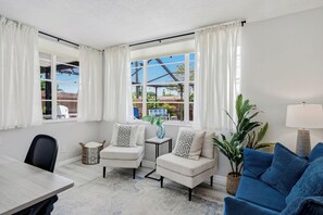 Ein Zimmer mit Esstisch, zwei Sesseln, einem blauen Sofa, einer Topfpflanze und einem fenster mit Vorhängen.