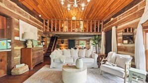 Interior de uma cabana de madeira com teto de madeira, sofá branco, mesa de centro redonda e escada que dá para um andar superior.