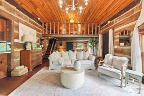 L’intérieur d’une cabane en rondins, avec un plafond en bois, un canapé blanc, une table basse ronde et un escalier menant à un niveau supérieur.