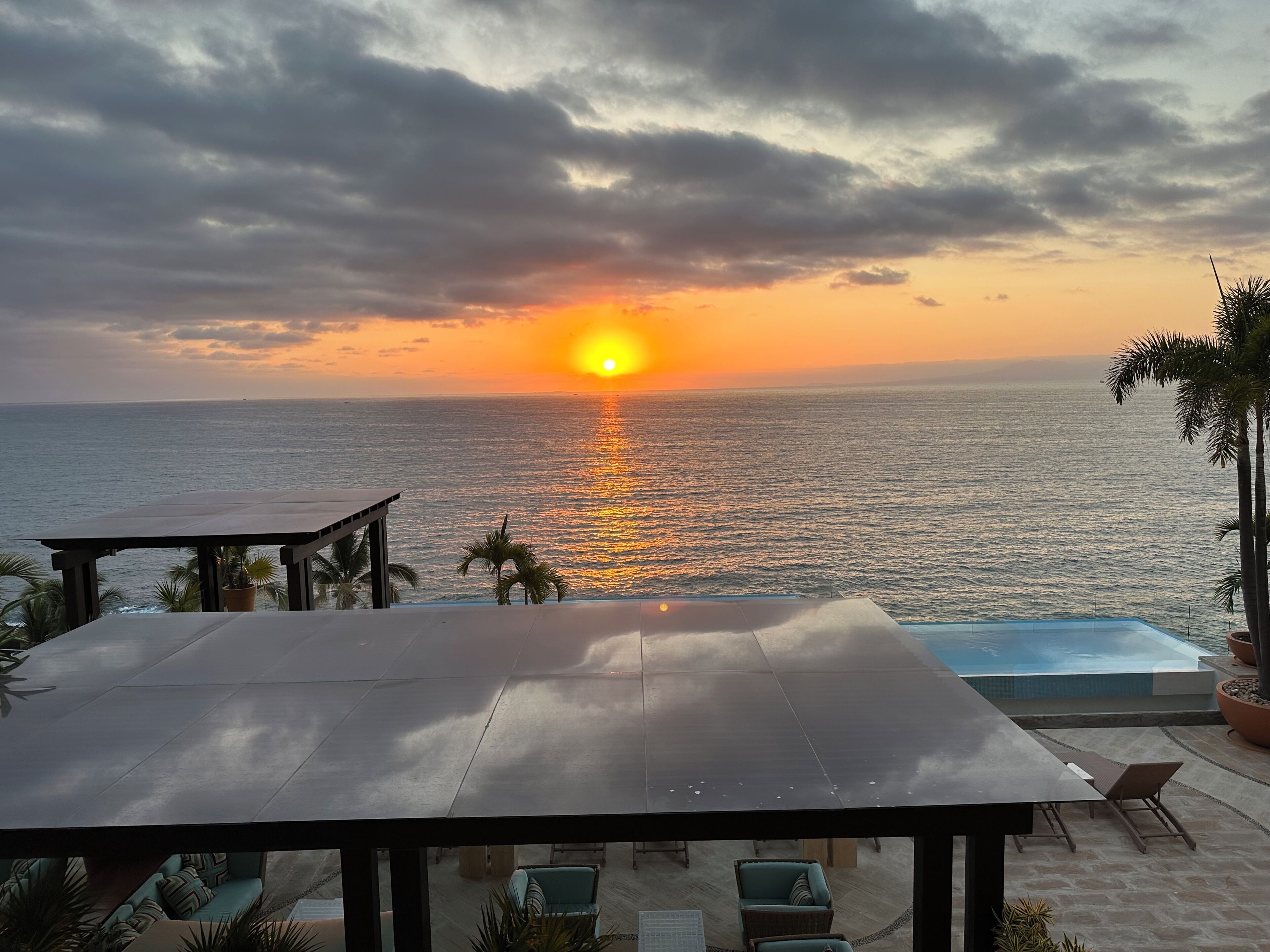 Een zonsondergang boven de oceaan, een zwembad en een overdekt terras met loungebedden.