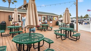 Une terrasse extérieure avec des tables vertes et des parasols, un bâtiment avec une enseigne indiquant « café » et « produits laitiers », et un drapeau en arrière-plan.