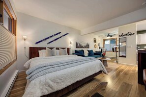Un dormitorio con una cama grande, un escritorio y una silla. Hay una ventana con persianas, una lámpara de pared y un cuadro.